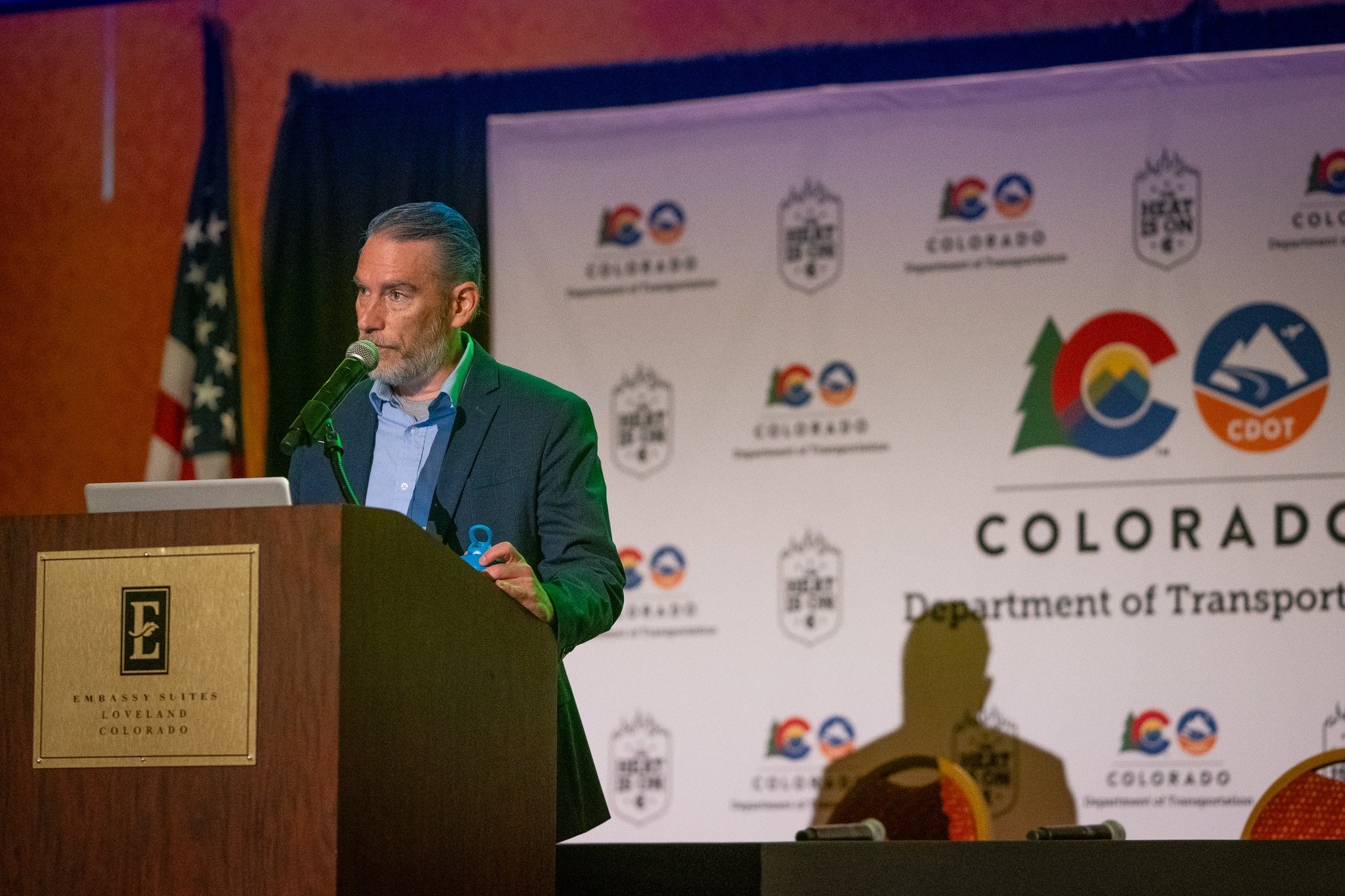 A speaker stands in front of a microphone at a podium. A backdrop with the Colorado Department of Transportation logo is behind him.